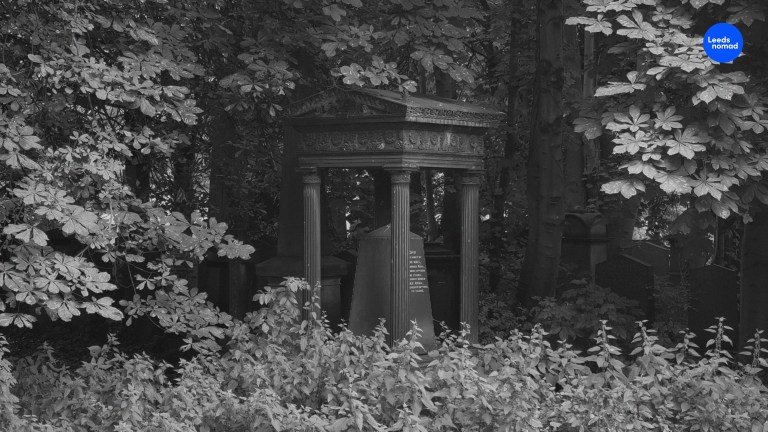 A black and white photo of a temple-like monument with four pillars under a canopy of trees surrounded by undergrowth.