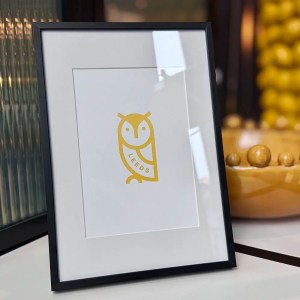 Gold printed Leeds Owl design on white paper in a black frame standing on a shelf.