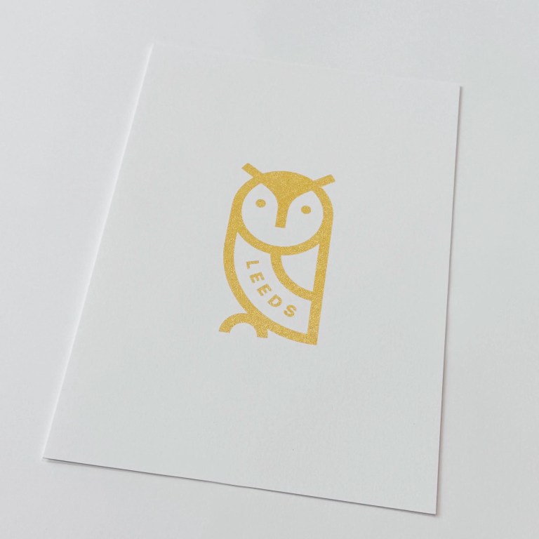 Gold printed Leeds Owl design on white paper in a black frame standing on a shelf.